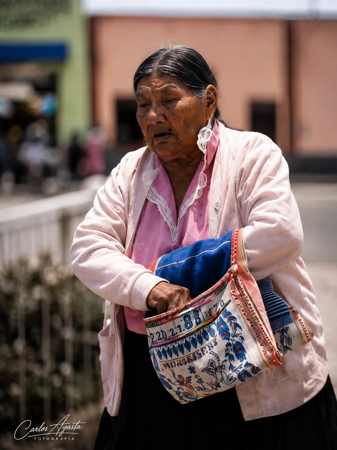 Mujer de pie lleva al hombro una alforja blanca con bordados de colores